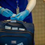 Mãos protegidas por luvas inspecionam etiquetas de bagagem durante procedimento de segurança em aeroporto.