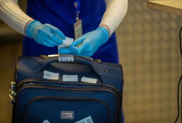 Mãos protegidas por luvas inspecionam etiquetas de bagagem durante procedimento de segurança em aeroporto.