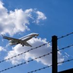 Avião se aproxima para pouso, visto contra um céu azul com nuvens, enquanto uma cerca de arame farpado aparece em primeiro plano.