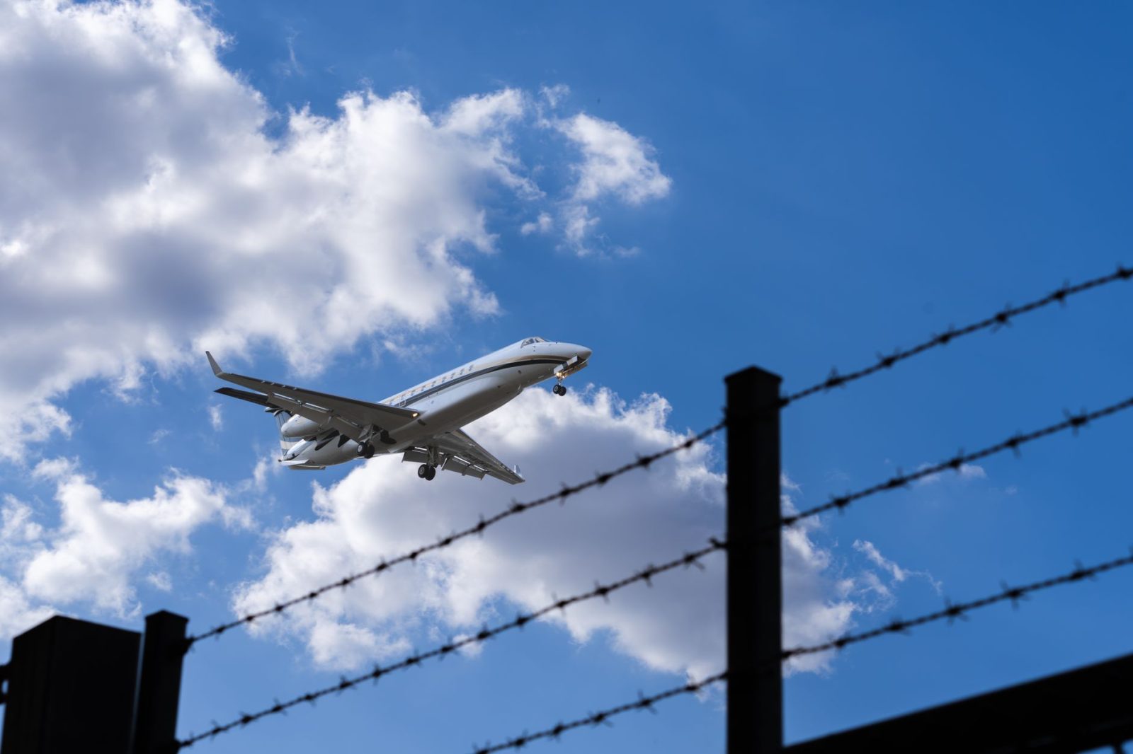 Avião se aproxima para pouso, visto contra um céu azul com nuvens, enquanto uma cerca de arame farpado aparece em primeiro plano.