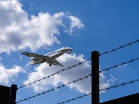 Avião se aproxima para pouso, visto contra um céu azul com nuvens, enquanto uma cerca de arame farpado aparece em primeiro plano.