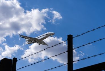 Avião se aproxima para pouso, visto contra um céu azul com nuvens, enquanto uma cerca de arame farpado aparece em primeiro plano.