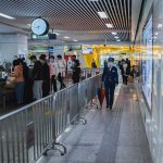 Passageiros passam pelo controle de segurança de um aeroporto, acompanhados por agentes e orientados por barreiras metálicas.