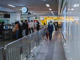 Passageiros passam pelo controle de segurança de um aeroporto, acompanhados por agentes e orientados por barreiras metálicas.
