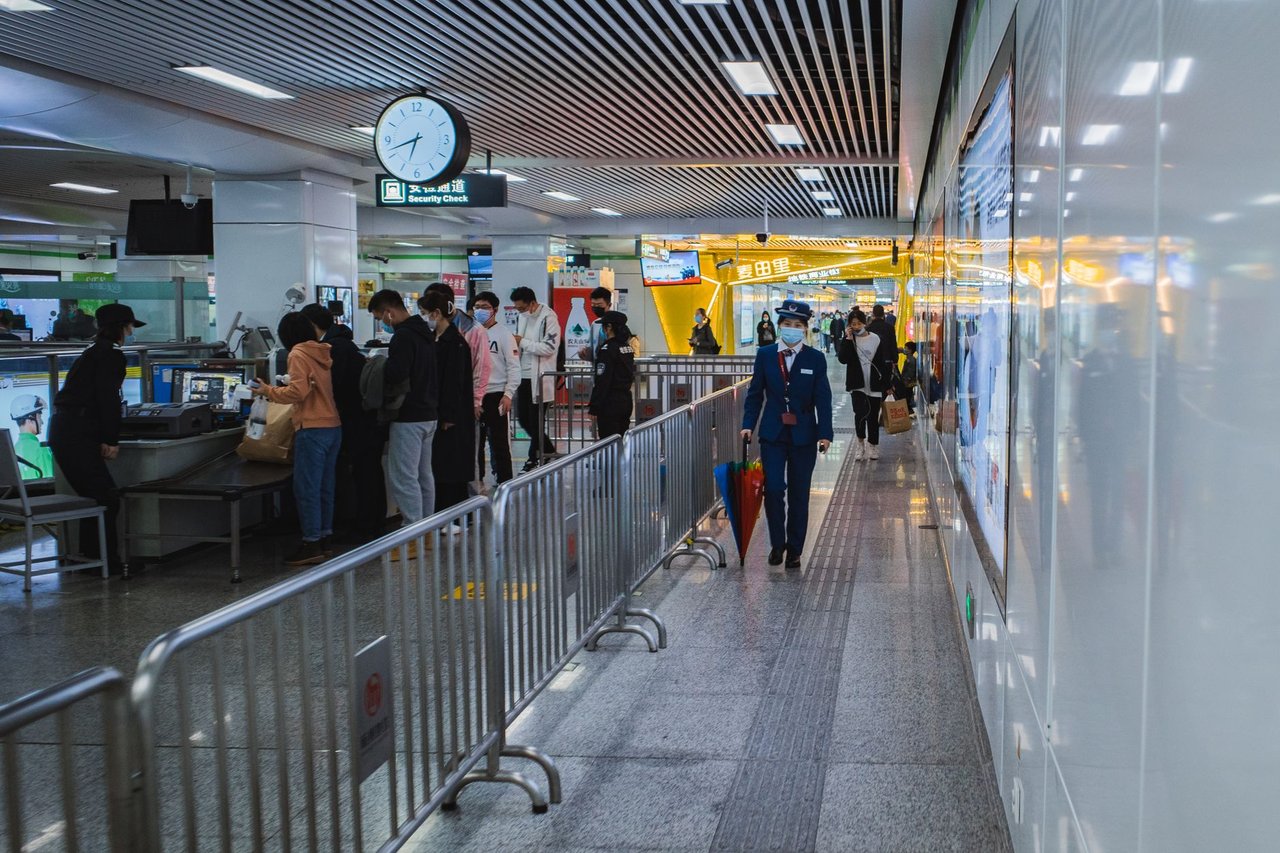 Passageiros passam pelo controle de segurança de um aeroporto, acompanhados por agentes e orientados por barreiras metálicas.