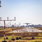 Avião pousa em uma pista de aeroporto, com a área urbana ao fundo