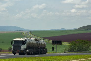 Caminhão de carga trafega por uma rodovia rural cercada por extensas áreas agrícolas.