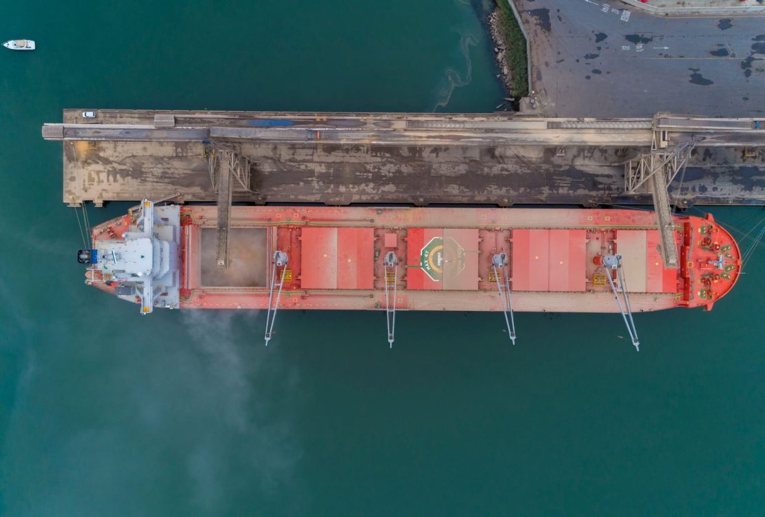Vista aérea de um navio cargueiro atracado no porto de São Francisco do Sul, com instalações portuárias e movimentação marítima ao redor.