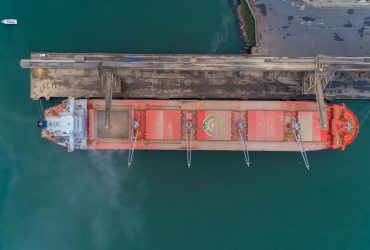 Vista aérea de um navio cargueiro atracado no porto de São Francisco do Sul, com instalações portuárias e movimentação marítima ao redor.
